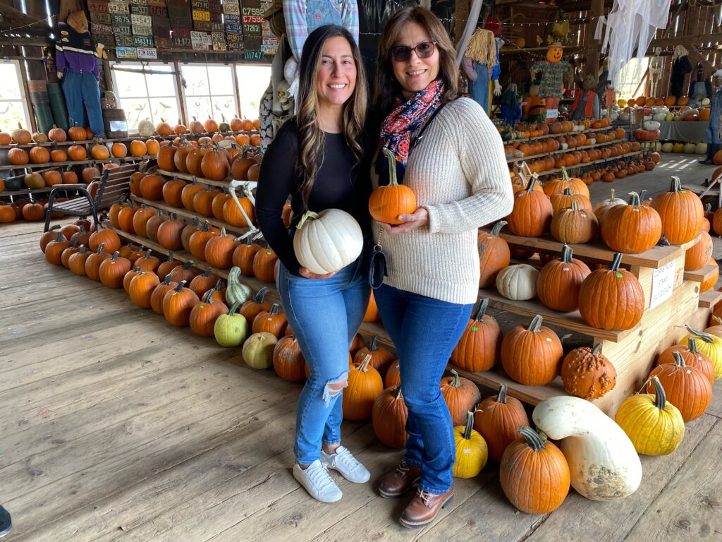 two women wearing sweaters holding pumpkins inside a barn full of different sized pumpkins