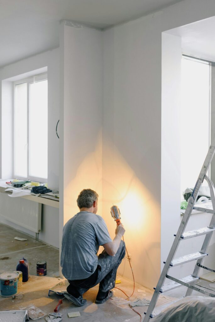 contractor working inside a home shining a light into a corner with a ladder beside him